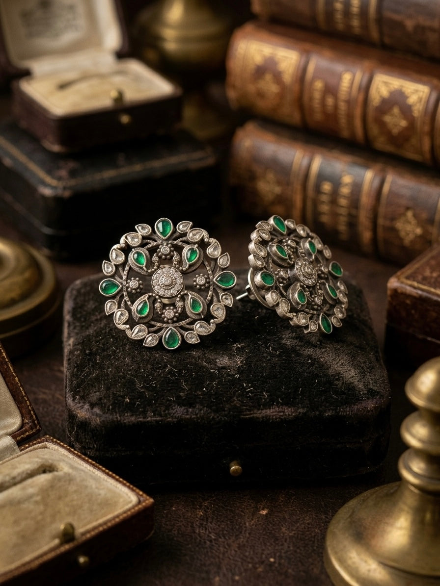 Pair of 925 sterling silver oxidised stud earrings with green emerald cabochons and polki uncut diamond accents in circular floral design, displayed on black velvet
