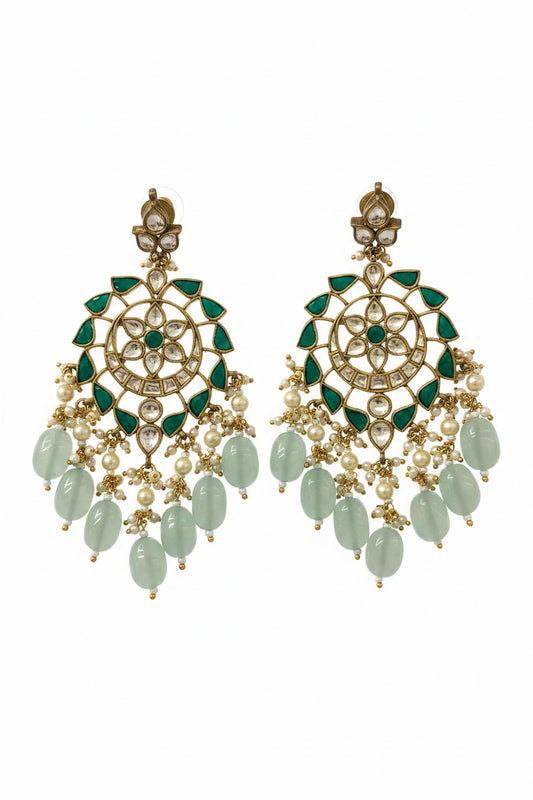 A pair of ornate gold-toned Indian chandelier earrings decorated with green teardrop crystals, white faux pearls, and light green stone drops, displayed on a textured gray fabric background.
